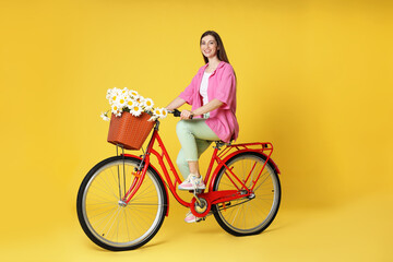 Smiling woman riding bicycle with basket of flowers against yellow background