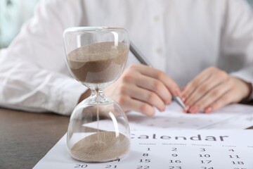 Deadline. Woman with calendar and pen at wooden table in office, focus on hourglass