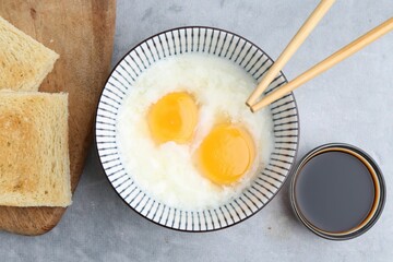Traditional asian breakfast with half-boiled eggs in bowl, soy sauce, bread and chopsticks on grey table, flat lay