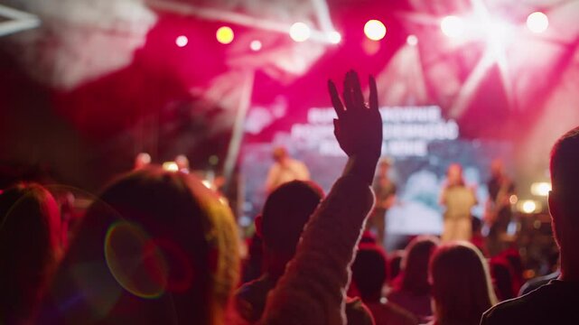 Person raises hand at outdoor concert or worship event, with colorful red stage lighting and blurred background. Ideal for music, celebration, church or festival atmosphere footage. 4K Backside shot