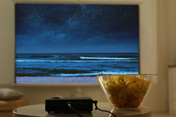Video projector and bowl with chips on wooden table. Night seascape on projection screen