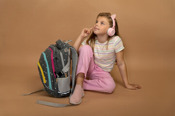 Little girl sitting next to school backpack wearing pink headphones and looking aside on beige...