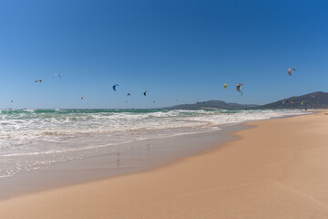 Kite surfers riding waves under a clear blue sky with colorful kites flying above the sea. Scenic summer beach view with mountains in the background in Tarifa, Spain