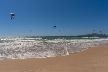 Kite surfers riding waves under a clear blue sky with colorful kites flying above the sea. Scenic...