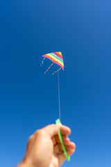 Close-up of a hand holding a reel while flying a colorful rainbow kite against a vibrant blue sky...