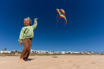Blond-haired boy flying a rainbow-colored kite on a sunny beach, smiling and enjoying a bright...
