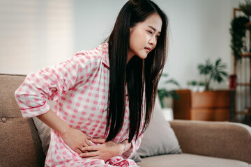 Young Asian woman suffering intense stomach ache abdominal pain sitting on comfortable couch...