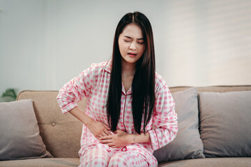 Young asian woman experiencing severe abdominal pain sitting sofa home health issue