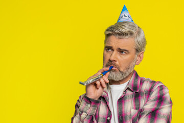 Mature man stands alone with party birthday cone hat and whistle blower showing dull spirit, disappointment. Middle-aged guy on yellow background feeling abandoned lonely, upset, tired, bored, unhappy