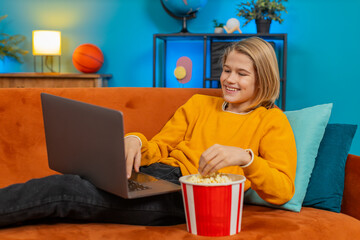 Caucasian teenager boy enjoys watching a movie on laptop at home. Chews popcorn, laughs heartily at funny scene, holds stomach while lying comfortably on sofa showing joy amusement in cozy living room
