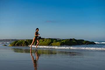 Young fit woman in green swimsuit running barefoot along wet beach, her reflection visible in the...