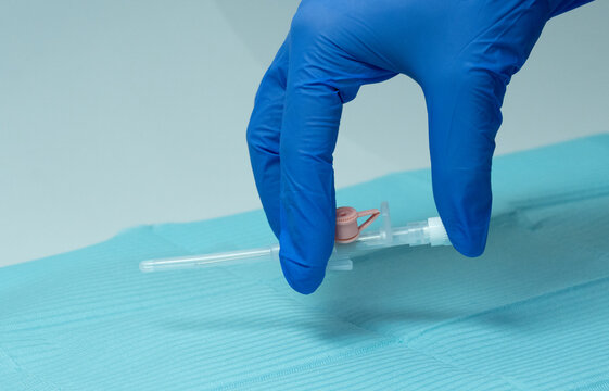 Gloved medical hand holding an IV cannula (venflon) in position just before insertion. Sterile setup with blue drape, showing preparation for intravenous access in clinical setting. - Powered by Adobe