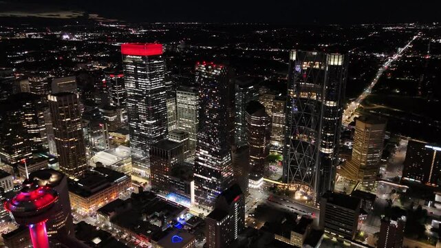 A cinematic night view of downtown Calgary, Alberta, showcasing the illuminated Bow Building, Calgary Tower, and TELUS Sky Tower under a dark sky. Captured in stunning detail, this urban skyline video