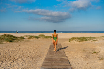 Woman in green swimsuit walking barefoot on a wooden beach path, heading toward the sea under a...