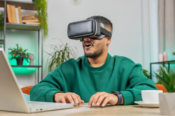 Indian man using virtual reality futuristic technology VR app headset helmet typing on virtual laptop keyboard, play simulation 3D 360 online video game at home office. Guy in goggles sitting at desk