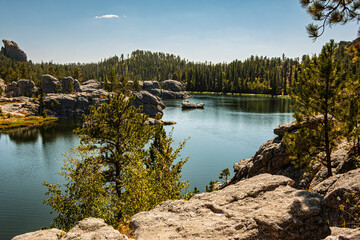 A scenic view of Sylvan Lake with granite boulders, evergreen trees, blue sky, and shimmering blue water. Custer State Park, Black Hills of South Dakota, USA.
