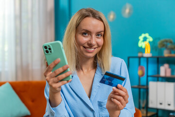 Young woman freelancer using credit bank card and smartphone phone while transferring money,...