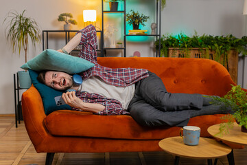 Repair work at neighbors. Irritated angry man lying on couch covers ears with cushions annoyed by...