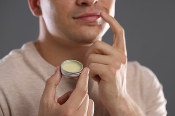 Young man applying lip balm onto his lips on grey background, closeup