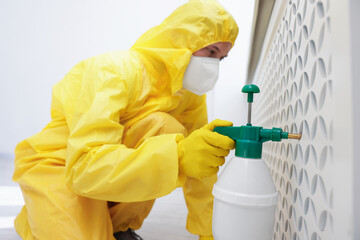 Pest control worker in protective suit spraying insecticide indoors, selective focus