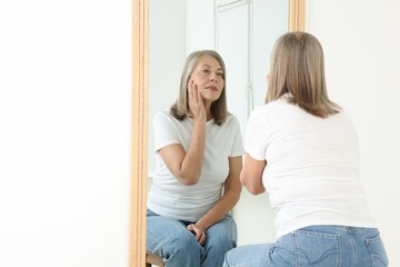 Beautiful senior woman looking at herself in mirror indoors