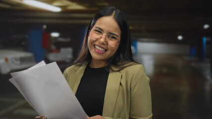 Young woman smiling reading documents in an indoor parking area wearing glasses and a beige blazer indicating a positive and confident attitude