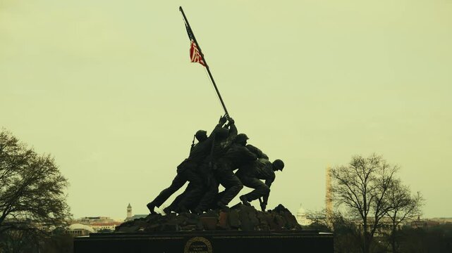 A silhouette of a statue of a group of soldiers raising a flag on top of a hill. The statue is set against a backdrop of a clear sky with a few clouds. The soldiers are depicted in a dynamic pose.