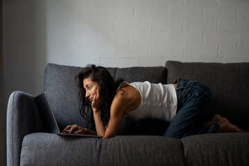 Focused female freelancer working on laptop while lying on sofa at home, immersed in creative...