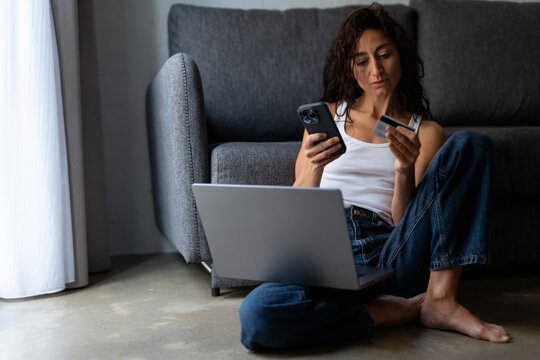 Serious young freelancer woman sitting on the floor at home with laptop, holding credit card and smartphone, checking online payment or banking account during remote work session.