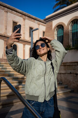 Fototapeta premium Stylish young woman in sunglasses taking selfie with smartphone on sunny day in urban setting.