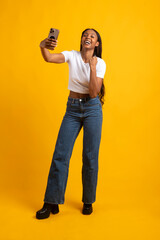 Excited young woman taking a selfie and celebrating success on yellow background