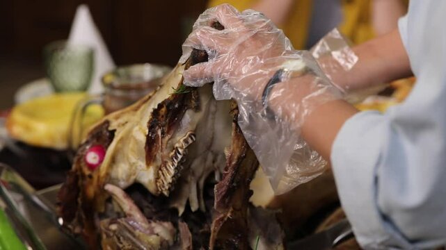 A gloved person carves a roasted lamb head, a traditional dish, serving the tender meat onto a plate. Ideal for culinary showcases, ethnic food blogs, and cultural dining experiences.