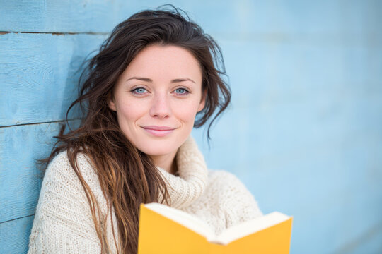 Young woman smiling and holding a book while leaning against a blue wall - Powered by Adobe