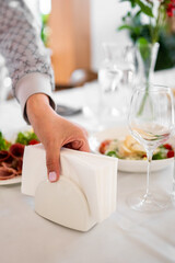Close-up of a hand placing a white napkin holder with napkins on a dining table set for a meal with food, drinks, and elegant tableware indoors.
