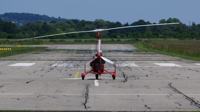 Red autogyro taxiing on the ground with spinning rotor blades under clear daylight
