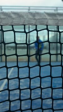 Through grid view of paddle tennis man playing on blue court outdoors. One caucasian padle player sportman hitting the ball. Racquet sport game Concept.