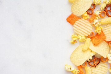 Salty snacks side border on a white marble background. Potato chips, popcorn, cheese puffs, pretzels. Above view.