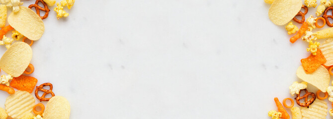 Salty snacks double border on a white marble banner background. Potato chips, popcorn, cheese puffs, pretzels. Overhead view.