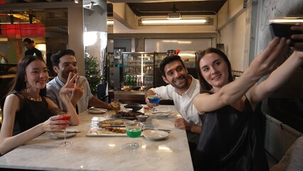 Woman taking sefie using smartphone with friends posing with special cocktail drinks surrounded by grill and fried meat special menu featuring unique chef table menu on table at restaurant. Vinosity.