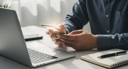 Working professional using smartphone beside laptop and notebook in bright modern office setting