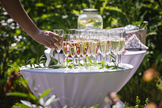 Hand of a person taking a glass from a table with champagne or sparkling white wine flutes with bubbles during an outdoor party under the sun