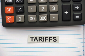 A grey strip of paper with the word Tariffs typed on it lying in an open notebook near a calculator.