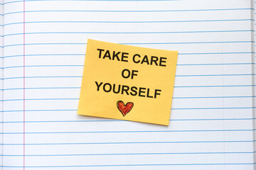 A yellow paper note with the words Take care of yourself on it lying in an open notebook. Close up.