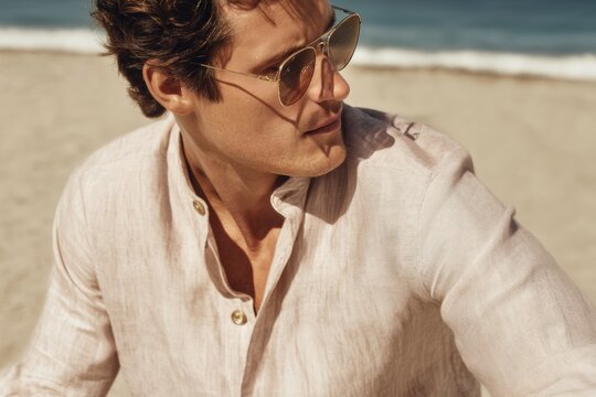 A man is seated on the sandy beach wearing a lightweight linen shirt and stylish sunglasses. He is enjoying the warm sunshine and the calming sound of the waves in the background