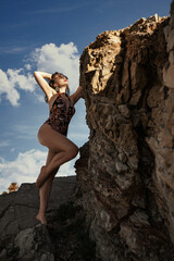 A woman in a colorful one-piece swimsuit and sunglasses poses against a sunlit, textured rock face, with a glimpse of water and clear sky.