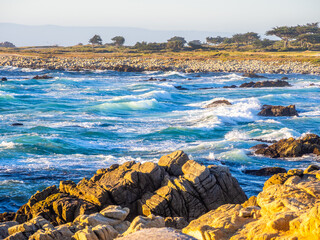 Waves crash on golden-lit rocks along the beginning of 17 Mile Drive