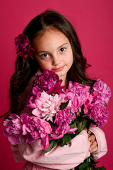 A young girl of Caucasian descent holds a vibrant bouquet of pink peonies against a bright pink...