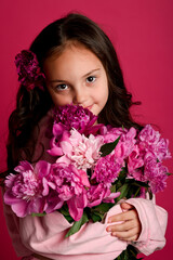 A young girl of Hispanic descent holds a bouquet of vibrant pink peonies against a vivid pink...