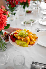 A hand holding a plate of fresh fruit over a festive table setting with elegant glassware, red flowers, and a bright, colorful atmosphere.