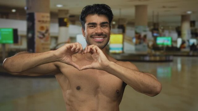 Beaming young shirtless man forms heart shape with his hands at crowded airport terminal checkin area; love.